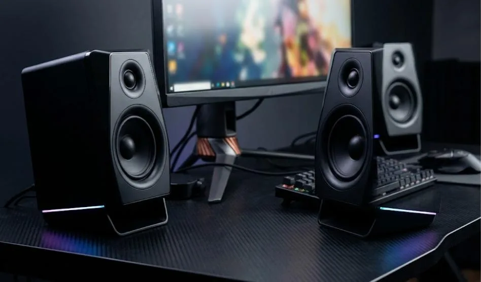 Pair of black desktop gaming speakers with RGB light strips beside a monitor on a dark desk